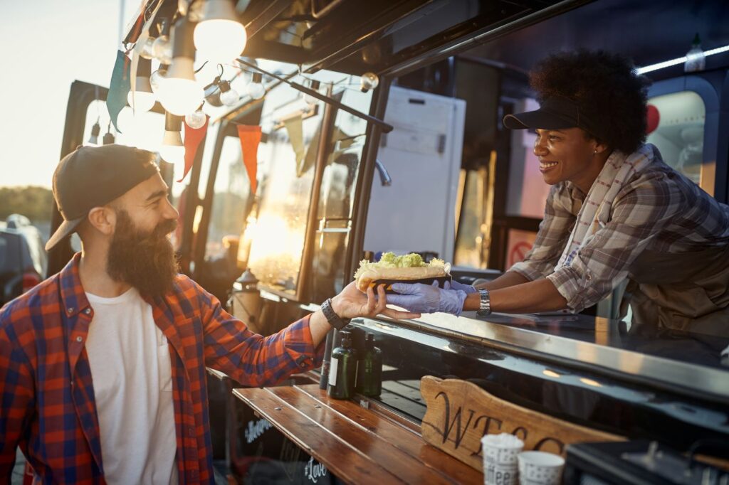 A smiling food truck owner cheerfully serves a delicious sandwich to her delighted customer.