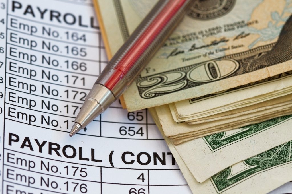 Stack of cash and a red pen lying on top of payroll paperwork. Represents the process of calculating payroll manually.